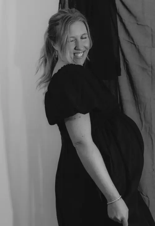 Lachende schwangere Frau im schwarzen Kleid hebt die Hand – lebendiger Studiomoment