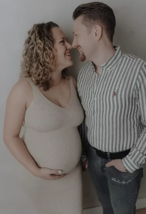 Schwangere Frau im beigen Kleid mit Partner, beide lächeln sich liebevoll an.